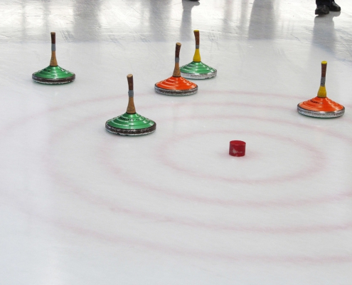 Beim Eisstockschießen liegt die Gaube im Mittelkreis. Eines der drei grünen Eisstöcke liegt am dichtesten am Ziel. Zwei orangene Stöcke kurz dahinter. Im Hintergrund sieht man die Füße der Spieler.