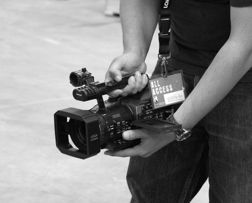 Schwarzweiß Foto eines Kameramannes, der seine Videokamera auf hüfthöhe hält und über den kleinen Kameramonitor etwas verfolgt. um den Hals hängt ein Schlüsselband mit der Karte "All Access".