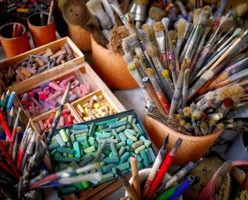 Ein sehr harmonisches, farbenfrohes Bild. Auf einem Tisch stehen diverse Dosen mit Pinseln, Stifte, Farben und Kreide. Es zeigt vorbereitende Maßnahmen für ein Puzzle Painting Event.